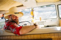 Man in cowboy hat sitting at drivers seat of retro van and looking at camera — Stock Photo