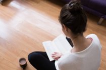 Вид зверху брюнетка дівчина сидить з ногами схрещеними на підлозі з чашкою кави і читання книги — стокове фото