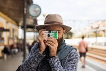 Retrato del hombre barbudo usando sombrero haciendo fotos con cámara de lomografía en la estación de tren - foto de stock