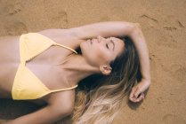 Вид зверху на молоду дівчину в жовтому купальнику, що лежить на піску пляжу з закритими очима . — стокове фото