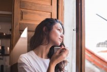 Стомлена молода жінка тримає чашку і дивиться на вікно — стокове фото