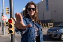 Jolie femme en lunettes de soleil debout au feu de circulation et arrêt gestuel — Photo de stock