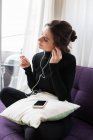 Retrato de chica morena sentada en el entrenador con smartphone y ajustando auriculares - foto de stock