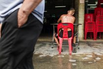 Вид сзади на человека без рубашки, сидящего на пластиковом стуле возле магазина на улице — стоковое фото