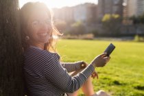 Усміхнена жінка сидить на траві зі смартфоном, дивлячись на камеру — стокове фото