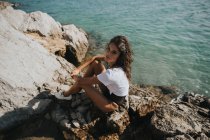 Portrait de fille brune assise sur une falaise sur la côte et regardant la caméra — Photo de stock