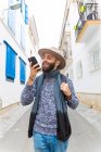 Bearded man in hat using voice search with smartphone while walking in street. — Stock Photo