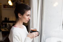 Vista laterale della donna bruna che posa con una tazza di cacao e guarda la finestra — Foto stock