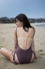 Back view of young girl posing in  swimming suit sitting on sand and looking over shoulder at camera — Stock Photo