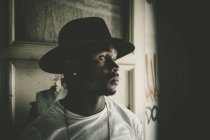 Hombre africano en sombrero en habitación oscura abandonada con graffiti en la pared. Mirando hacia otro lado . - foto de stock