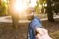 Bearded man in denim jacket and cap holding hand and gesturing Follow me in sunlight. — Stock Photo