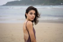 Slim ragazza bruna in costume da bagno guardando oltre la spalla alla fotocamera sulla spiaggia — Foto stock