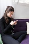 Portrait of brunette girl in earphones sitting on coach and browsing smartphone — Stock Photo