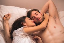 Mujer joven abrazando y mirando al novio soñoliento estirándose en la cama - foto de stock