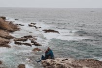Visão traseira do casal romântico sentado na costa rochosa e admirando a paisagem marinha — Fotografia de Stock