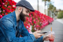 Seitenansicht einer bärtigen Person, die beim Smoothie-Trinken am Smartphone sitzt und surft. — Stockfoto