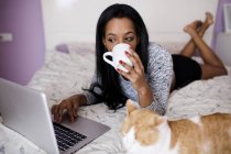 Mädchen trinkt Kaffee, während sie im Bett am Laptop surft — Stockfoto
