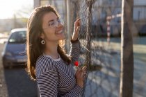 Pretty girl smiling and leaning at metal grid on city street. — Stock Photo