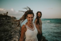 Portrait of happy woman with dreadlocks holding male hand and looking at camera on background of tropical beach. — Stock Photo