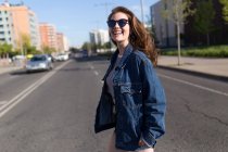 Joyeux jeune femme en lunettes de soleil regardant la caméra tout en traversant la route . — Photo de stock