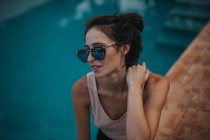 Портрет стильної брюнетки в дзеркальних сонцезахисних окулярах, що сидять на краю басейну — стокове фото