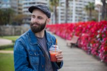 Retrato de homem barbudo posando com smoothie no parque e olhando para longe — Fotografia de Stock