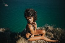 Vue grand angle de la femme brune posant les yeux fermés sur la roche au-dessus de la surface de l'océan — Photo de stock