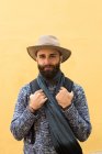 Portrait of bearded man in hat posing with backpack over yellow wall and looking at camera — Stock Photo