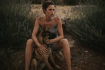 Retrato de morena em vestido sentado em pedra e abraçando cão pastor no campo — Fotografia de Stock