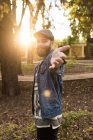 Portrait of bearded man in denim clothing smiling and outstretching hand at camera on background of park in sunlight. — Stock Photo