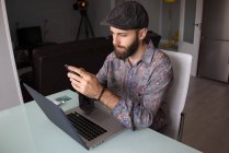 Портрет бородатого чоловіка, який сидить за столом з ноутбуком і використовує смартфон — стокове фото