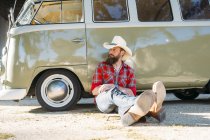 Hombre barbudo con sombrero de vaquero sentado en el suelo y apoyado en la furgoneta retro y mirando hacia otro lado - foto de stock