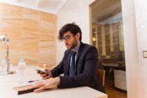 Jovem empresário de terno sentado na sala de reuniões e smartphone de navegação . — Fotografia de Stock