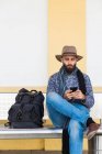 Бородатый мужчина с рюкзаком сидит на скамейке и слушает музыку с наушниками во время просмотра смартфона — стоковое фото