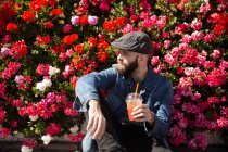 Vista frontal del hombre barbudo sentado cerca de las flores y sosteniendo batido durante mirar hacia otro lado . - foto de stock