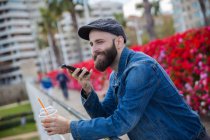 Vista laterale dell'uomo barbuto sorridente che tiene in mano tazza di bevanda e voce alla ricerca con smartphone . — Foto stock