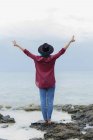 Rear view of stylish incognito girl in black hat standing on rocks at seaside and showing peace gesture both hands.  Copyspace. — Stock Photo