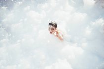 Cheerful kid playing in white foam — Stock Photo