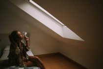 Junge brünette Frau raucht Zigarette, während sie auf dem Bett gegen das Dachbodenfenster sitzt — Stockfoto