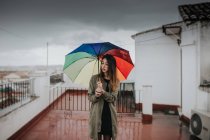 Young brunette standing on building roof with umbrella colored rainbow. Copyspace. — Stock Photo