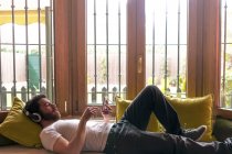 Молодий чоловік слухає музику з планшетом, розслабленим вдома з вікном, освітленим теплим світлом сонця — стокове фото