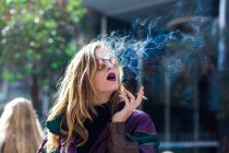 Молода стильна жінка в сонцезахисних окулярах чуттєво курить на розмитому фоні вулиці . — стокове фото