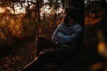 Бородатый человек сидит на земле за деревом в осеннем лесу — стоковое фото
