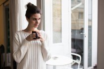 Portrait of cute brunette girl posing with cup near window and looking aside — Stock Photo