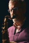 Зрілий чоловік в окулярах грає на саксофоні з закритими очима на чорному тлі — стокове фото
