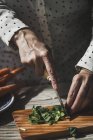 Vista da vicino delle mani affettare foglie di basilico con coltello su tavola di legno — Foto stock