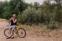 Портрет старшого чоловіка, що спирається на велосипед і розмовляє через смартфон у парку — стокове фото