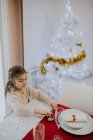 Високий кут зору маленької дівчинки, що подає ложку за різдвяним столом біля білої ялинки . — стокове фото