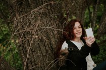 Улыбающаяся девушка делает селфи за деревом в лесу — стоковое фото