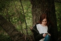 Chica jengibre sentado junto al árbol y el teléfono inteligente de navegación - foto de stock